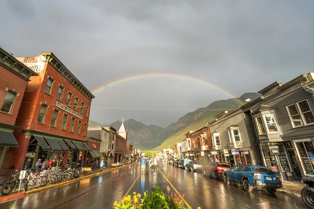 A vibrant street scene featuring historic buildings, a rainbow overhead, wet pavement from recent rain, and a mountain backdrop. People are seen exploring local shops, contributing to a communal, lively, and walkable neighborhood vibe.