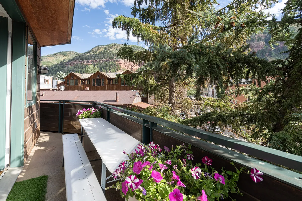 This inviting exterior shot showcases a tranquil terrace adorned with vibrant pink flower planters and comfortable wooden bench seating, surrounded by lush trees and set against a dramatic mountain backdrop. The scene hints at relaxing mornings, sunset gatherings, and panoramic alpine views, all within the heart of Telluride.