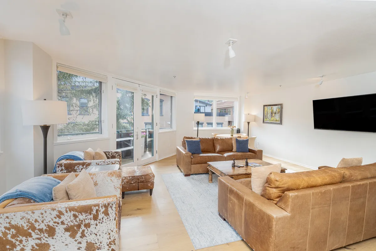 A spacious living area featuring a large brown leather sofa, accent chairs, a coffee table, and wide windows that frame sweeping mountain and town views. A television anchors the space, while the seating arrangement encourages both conversation and relaxation. The room is flooded with natural light and exudes a sense of comfort and warmth.