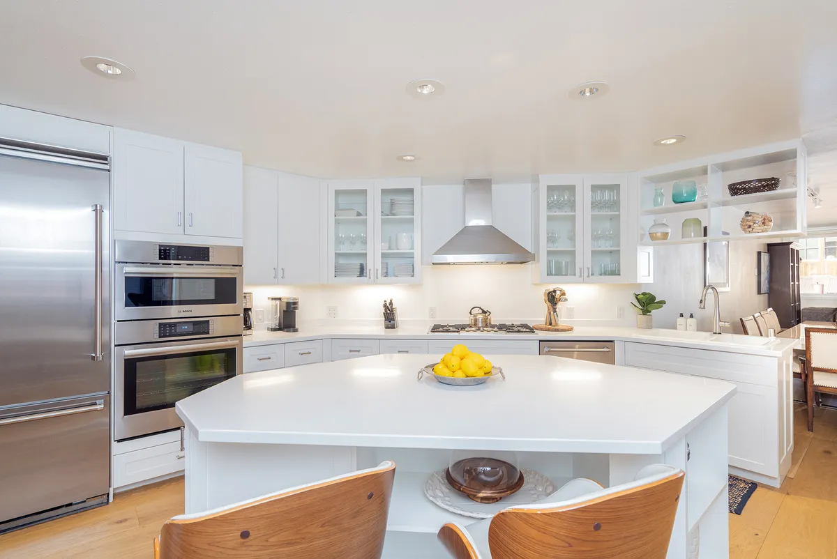 A warmly lit chef’s kitchen featuring a spacious island, gleaming stainless steel Viking appliances, glass-front cabinets, and a colorful fruit bowl. The kitchen setup invites both meal preparation and social interaction, with modern finishes and an inviting layout.