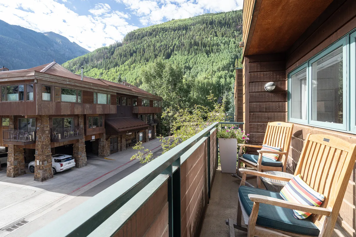 A tranquil balcony scene with comfortable seating, overlooking stunning mountain views and lush greenery. The adjacent building’s architecture hints at a communal living atmosphere, while the setting is perfect for small gatherings, relaxing with friends, or sharing a quiet meal outdoors.