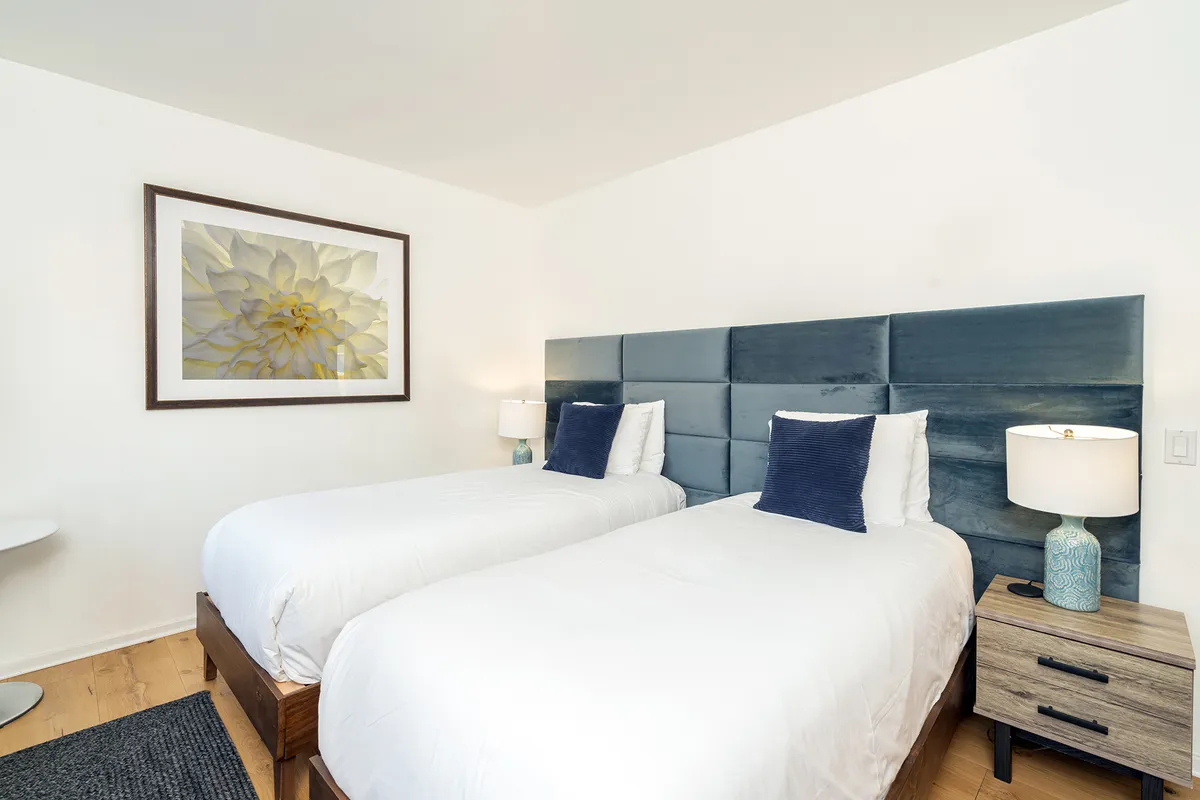 A cozy and serene second bedroom setup with two twin beds, framed flower artwork above the beds, and a nightstand with a lamp in between. The space feels peaceful, providing a flexible arrangement for families or friends.