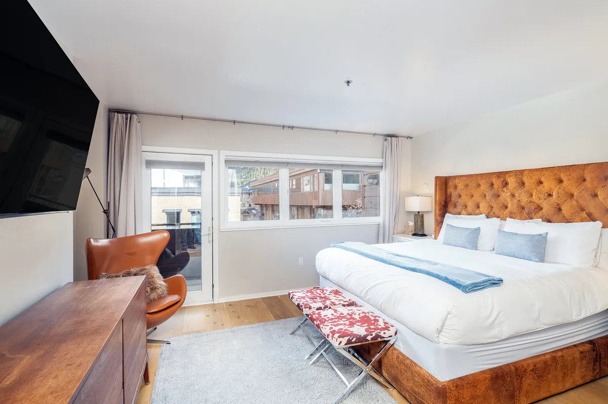 This image features a spacious primary bedroom with a large tufted headboard king bed, a modern TV, a comfortable armchair, a wooden dresser, and a stylish accent bench. Natural light pours in through the window, creating a serene atmosphere ideal for relaxation and morning routines.