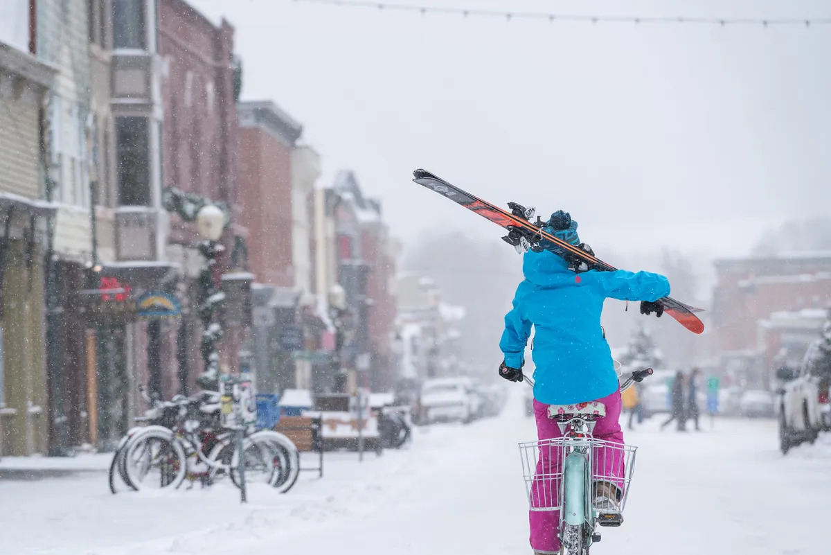 A person rides a bicycle with skis attached, passing along a snow-covered street lined with colorful buildings. The scene is bustling with winter activity, suggesting proximity to both the slopes and local amenities.