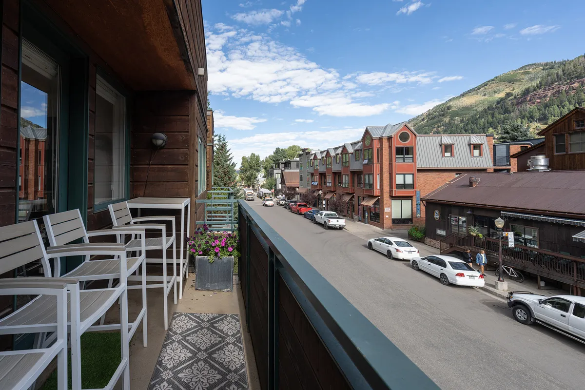 A tranquil balcony features comfortable seating, flower pots, and a view over a bustling street with a stunning mountain backdrop. The scene balances relaxation with a direct connection to both town life and nature.