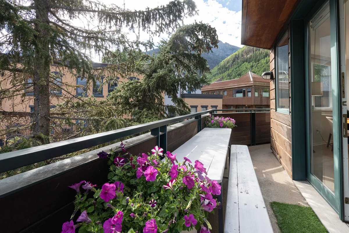 A tranquil wrap-around terrace adorned with vibrant purple flowers, a wooden balcony railing, and comfortable seating that overlooks a stunning mountain landscape. The ambiance is peaceful yet lively, ideal for enjoying coffee at sunrise or toasting the sunset with friends while taking in the alpine scenery.