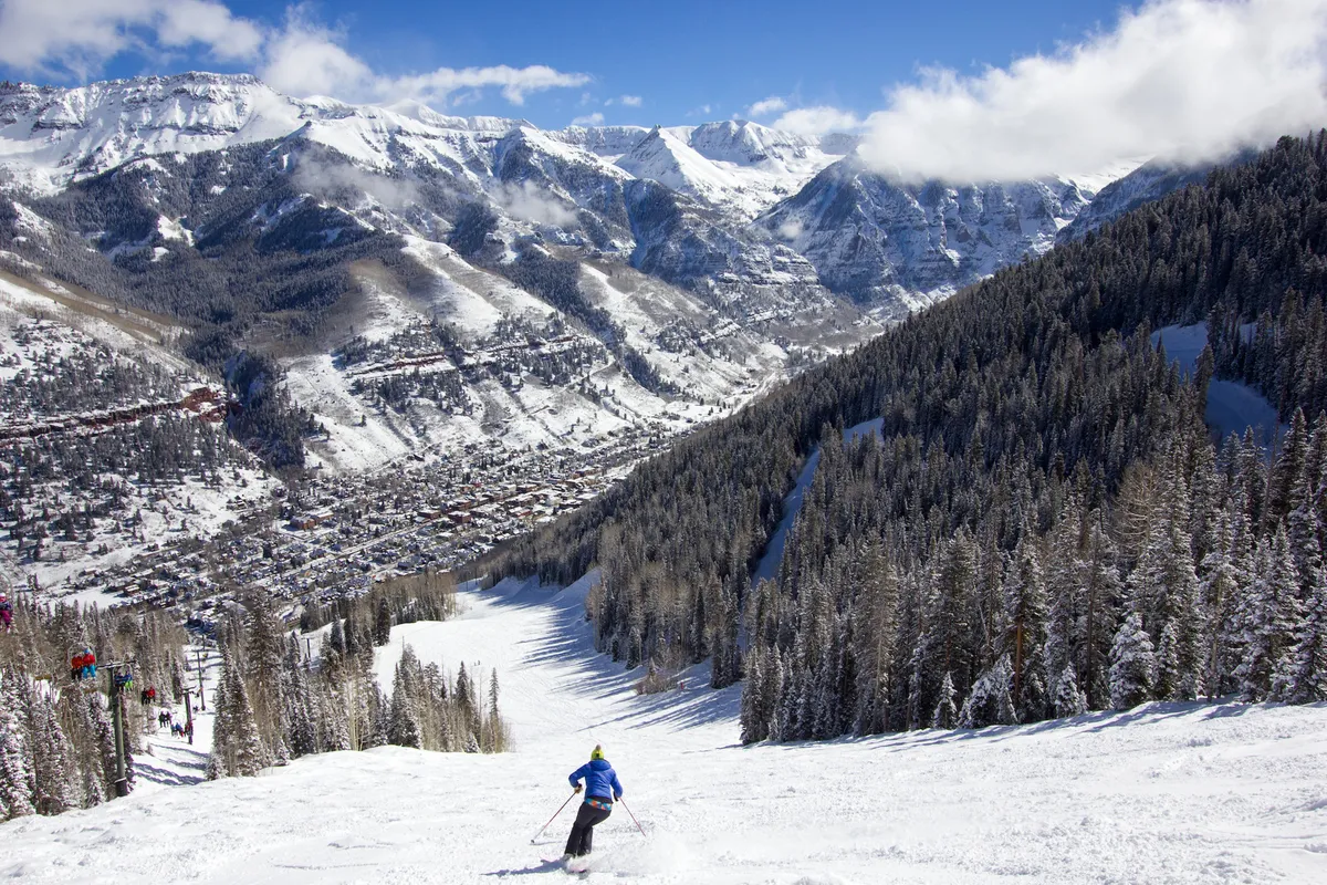 A skier carves down a snowy mountain slope, with pine trees, distant peaks, and the town visible below. This action-filled image captures the thrill of winter sports and the proximity of world-class skiing.