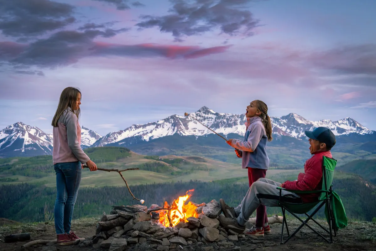 Children gathered around a campfire, roasting marshmallows with a scenic mountain backdrop. The joyful, nostalgic mood captures the essence of outdoor family fun and creating special memories together.