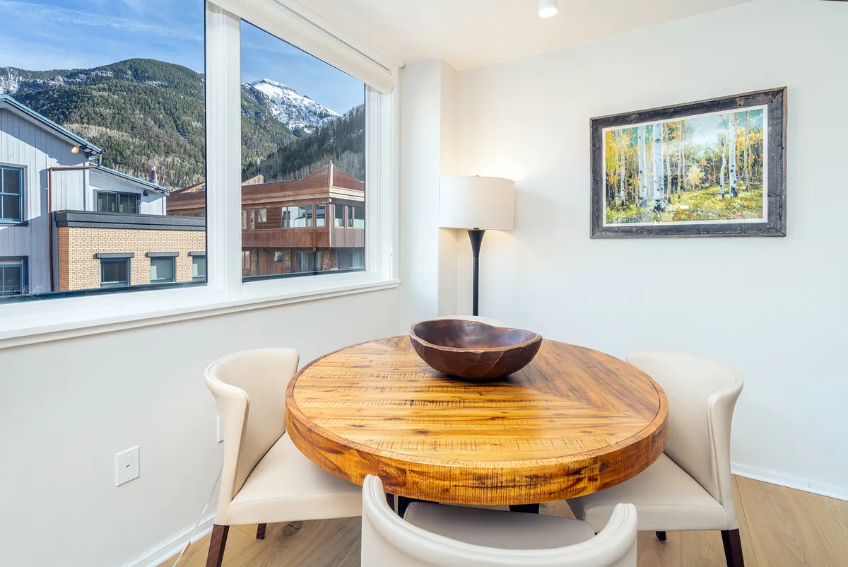 A round wooden game or dining table set by large windows with a mountain view. The table is accompanied by seating, a floor lamp, and aspen tree artwork, creating an inviting niche for conversation, games, or enjoying coffee while taking in the sunrise over the peaks.