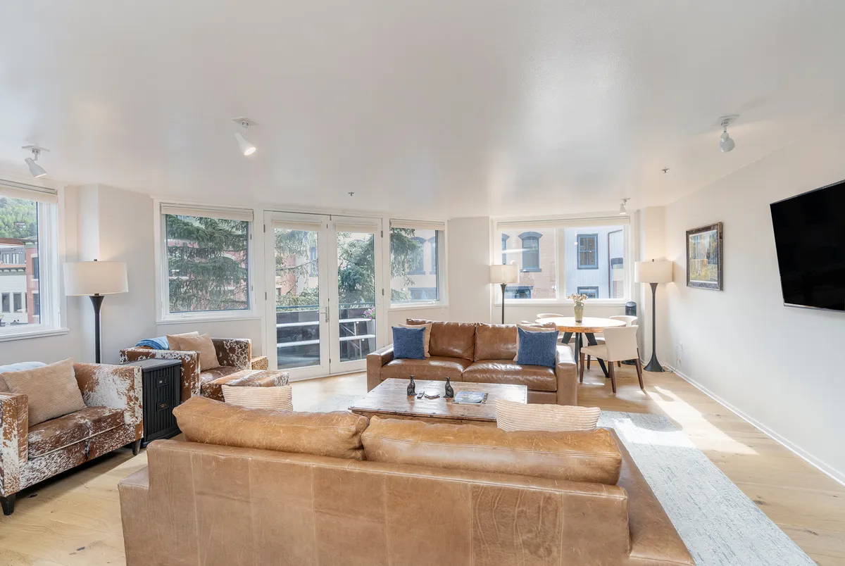 A spacious living and dining area is furnished with a brown leather sofa, comfortable armchairs, a dining table, and large windows that let in plenty of natural light. The space is ideal for relaxing, entertaining, or enjoying a quiet meal at home.
