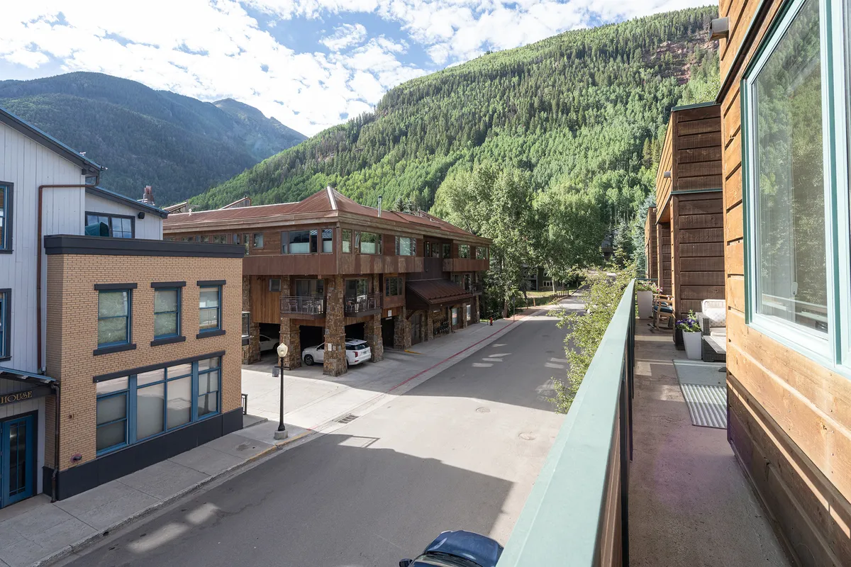 A lively street scene in the heart of town, with modern buildings set against a backdrop of mountains, a tree-lined street, and hints of pedestrian activity. The environment looks safe and vibrant, perfect for families to walk, bike, and explore local shops or attend community events.