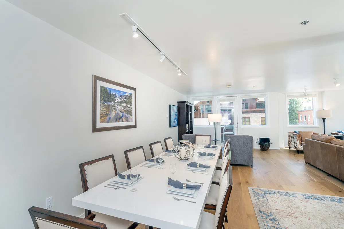 A contemporary dining area with a large table set for a group, stylish artwork on the wall, natural light streaming through windows, and a sofa in the background. The space is arranged for lively meals, games, or social gatherings, radiating a warm and inviting atmosphere.