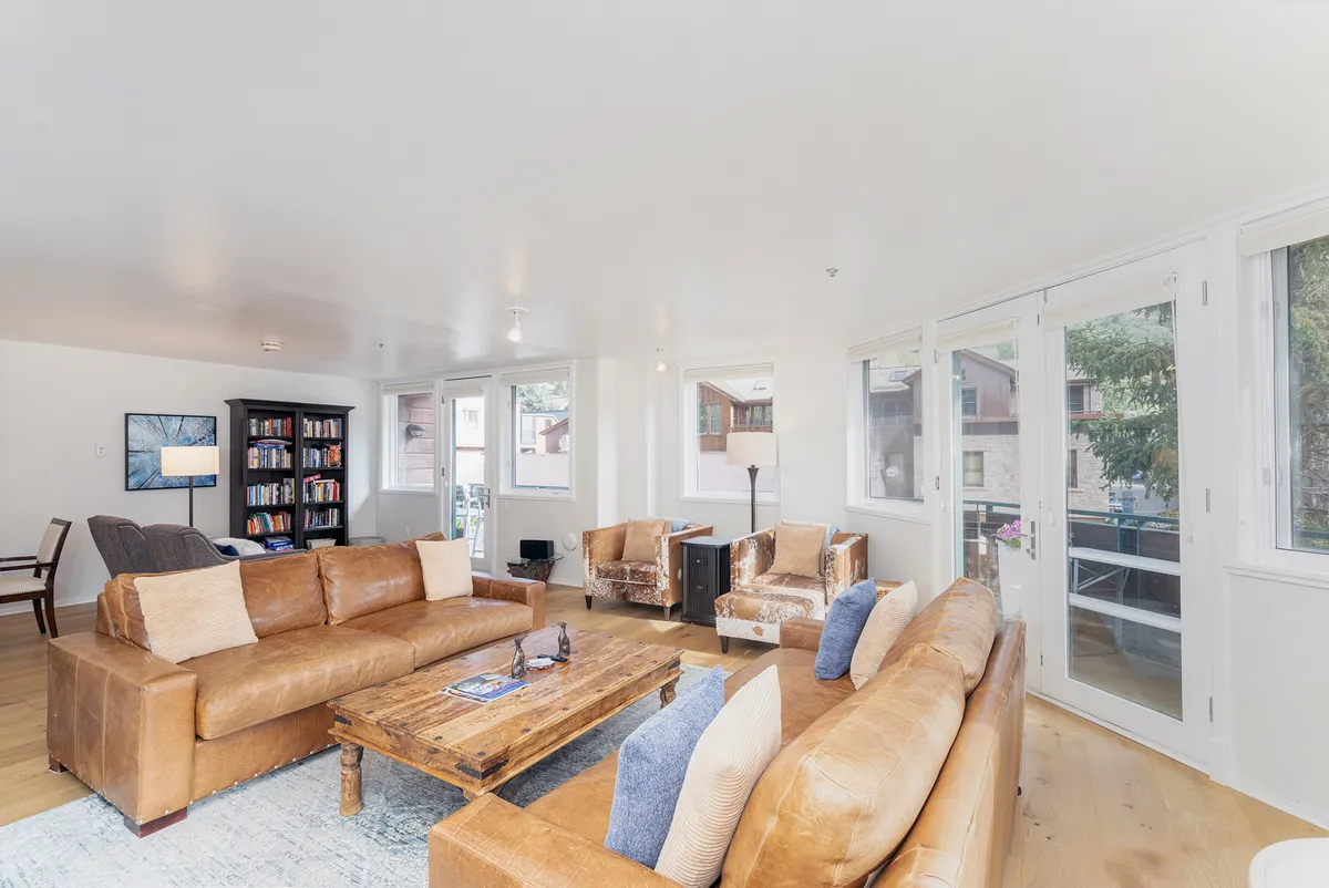 A welcoming living room with brown leather sofas, a wooden coffee table, a bookshelf filled with books, and expansive large windows that flood the space with natural light. The windows offer glimpses of the surrounding mountain landscape. The overall mood is cozy, social, and ideal for both relaxation and gathering with friends or family.